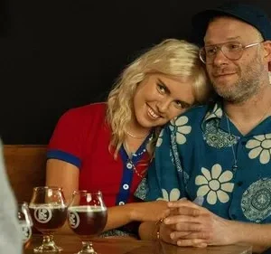 Casal sorridente num bar. Mulher loira encosta no ombro de homem barbudo de camisa florida. Bebem cervejas, clima afetuoso.