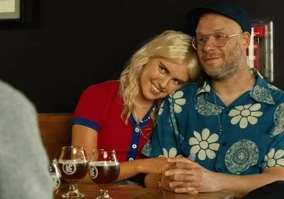 Casal sorridente num bar. Mulher loira encosta no ombro de homem barbudo de camisa florida. Bebem cervejas, clima afetuoso.
