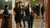 Duas mulheres caminham em um escritório movimentado. Uma em vestido preto sorri; a outra, de terno e com bolsas, parece tensa.