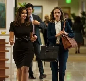 Duas mulheres caminham em um escritório movimentado. Uma em vestido preto sorri; a outra, de terno e com bolsas, parece tensa.