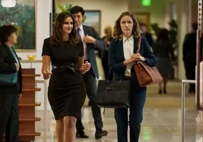 Duas mulheres caminham em um escritório movimentado. Uma em vestido preto sorri; a outra, de terno e com bolsas, parece tensa.