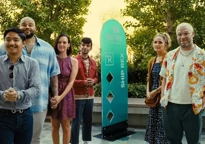 Seis pessoas sorridentes, de diversas etnias, posam em um ambiente externo ensolarado com banners coloridos. Clima alegre e casual.