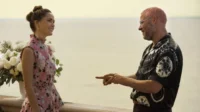 Homem de cabelo rosa e camisa tie-dye aponta para mulher em vestido floral rosa, sorrindo. Estão em varanda com flores, vista para o mar.