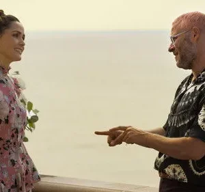 Homem de cabelo rosa e camisa tie-dye aponta para mulher em vestido floral rosa, sorrindo. Estão em varanda com flores, vista para o mar.
