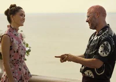 Homem de cabelo rosa e camisa tie-dye aponta para mulher em vestido floral rosa, sorrindo. Estão em varanda com flores, vista para o mar.