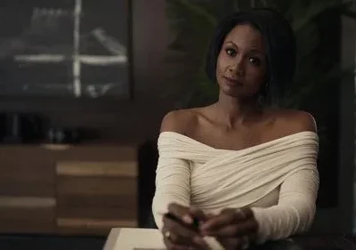 Mulher de pele escura com cabelo curto e vestido creme de ombros caídos em ambiente moderno e elegante. Olhar direto.
