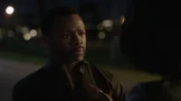 Homem negro de barba e camisa escura olha intensamente para outro indivíduo. Cenário noturno com luzes urbanas desfocadas.