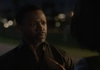 Homem negro de barba e camisa escura olha intensamente para outro indivíduo. Cenário noturno com luzes urbanas desfocadas.