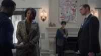 Três pessoas sérias em uma sala luxuosa. Um homem de costas, uma mulher de casaco elegante e um homem de terno olham-se. Uma pessoa com óculos anota.