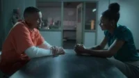 Homem de macacão laranja sorri para mulher séria com mãos unidas. Eles estão numa sala de interrogatório escura.