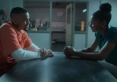 Homem de macacão laranja sorri para mulher séria com mãos unidas. Eles estão numa sala de interrogatório escura.