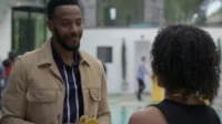 Homem negro sorridente com jaqueta bege e polo conversa com mulher de cabelo cacheado, de costas, em festa ao ar livre com piscina.