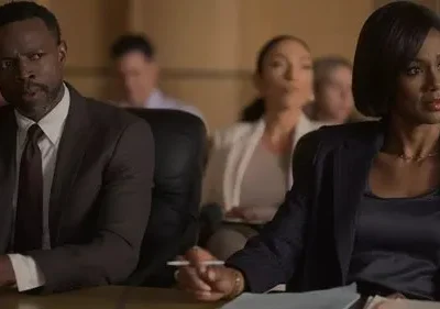 Um homem e uma mulher de terno sentados em uma mesa formal. Ele com mãos postas, ela com caneta e olhar focado. Clima sério e atento.