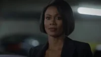 Mulher de pele escura, cabelo curto e casaco escuro, com expressão séria. Fundo urbano escuro com luzes borradas.