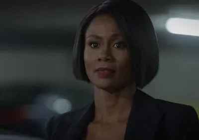 Mulher de pele escura, cabelo curto e casaco escuro, com expressão séria. Fundo urbano escuro com luzes borradas.