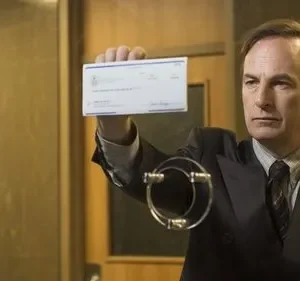 Homem de terno, com olhar sério, ergue um cheque branco em um balcão. O ambiente é um escritório formal.