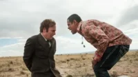 Em um deserto, um homem de terno, amedrontado, é confrontado por outro homem que se inclina, com camisa colorida e postura ameaçadora.
