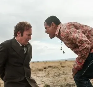 Em um deserto, um homem de terno, amedrontado, é confrontado por outro homem que se inclina, com camisa colorida e postura ameaçadora.