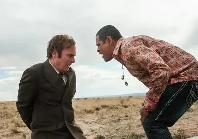 Em um deserto, um homem de terno, amedrontado, é confrontado por outro homem que se inclina, com camisa colorida e postura ameaçadora.