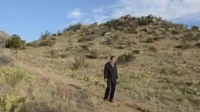 Homem de terno parado numa trilha, em paisagem desértica com colinas, cactos e céu azul. Ambiente vasto e isolado.