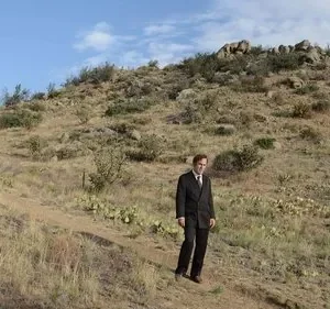 Homem de terno parado numa trilha, em paisagem desértica com colinas, cactos e céu azul. Ambiente vasto e isolado.