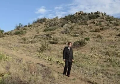 Homem de terno parado numa trilha, em paisagem desértica com colinas, cactos e céu azul. Ambiente vasto e isolado.