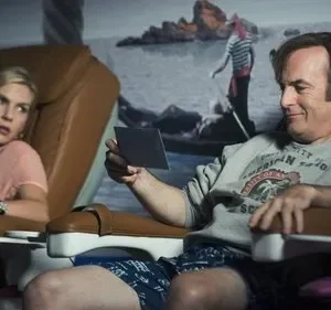 Homem de moletom sorri para foto em cadeira de salão; mulher loira ao lado parece tensa. Fundo com mural de praia.