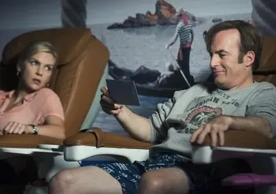 Homem de moletom sorri para foto em cadeira de salão; mulher loira ao lado parece tensa. Fundo com mural de praia.