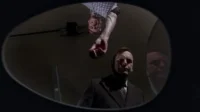 Vista de baixo, um homem de terno olha intensamente enquanto outro, invertido, oferece um objeto. A imagem é escura e misteriosa.