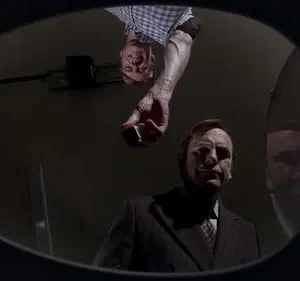 Vista de baixo, um homem de terno olha intensamente enquanto outro, invertido, oferece um objeto. A imagem é escura e misteriosa.