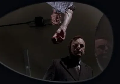 Vista de baixo, um homem de terno olha intensamente enquanto outro, invertido, oferece um objeto. A imagem é escura e misteriosa.