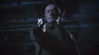 Homem careca e barbudo com casaco, olhar sério, aponta arma brilhante com luva em cenário escuro. Clima sombrio e tenso.