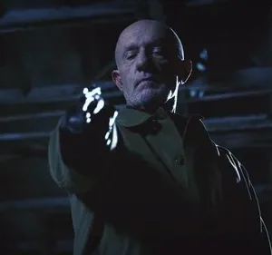 Homem careca e barbudo com casaco, olhar sério, aponta arma brilhante com luva em cenário escuro. Clima sombrio e tenso.