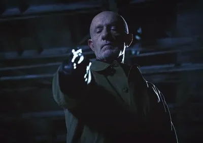 Homem careca e barbudo com casaco, olhar sério, aponta arma brilhante com luva em cenário escuro. Clima sombrio e tenso.