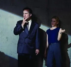 Homem de terno fumando e mulher séria em garagem escura. A luz cria sombras dramáticas na parede de concreto.