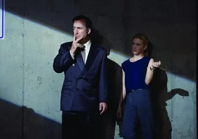 Homem de terno fumando e mulher séria em garagem escura. A luz cria sombras dramáticas na parede de concreto.