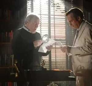 Em um escritório, um homem de suéter usa lupa para examinar papéis com outro de terno claro. Estantes e luz intensa pela janela criam clima sério.