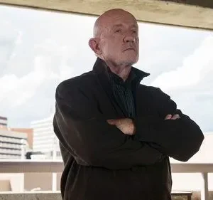 Homem careca e idoso, braços cruzados, olhar sério, contra um céu nublado e edifícios ao fundo. Clima tenso.