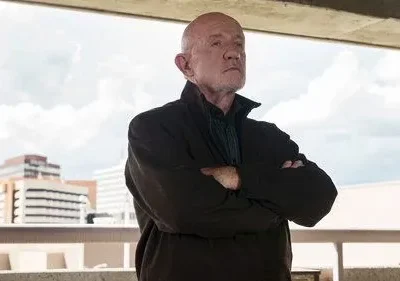 Homem careca e idoso, braços cruzados, olhar sério, contra um céu nublado e edifícios ao fundo. Clima tenso.