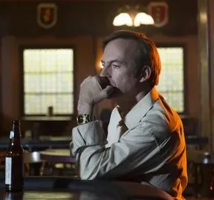 Homem pensativo com a mão no queixo sentado em um bar escuro. Há uma garrafa de cerveja no balcão e um letreiro neon ao fundo.