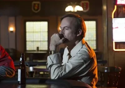 Homem pensativo com a mão no queixo sentado em um bar escuro. Há uma garrafa de cerveja no balcão e um letreiro neon ao fundo.