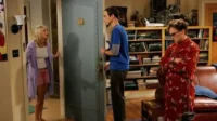 Uma mulher de pijama surpresa na porta, um homem sério e outro de roupão resmungão numa sala com livros.