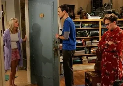 Uma mulher de pijama surpresa na porta, um homem sério e outro de roupão resmungão numa sala com livros.