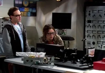 Dois cientistas em um laboratório cheio de equipamentos. Um homem de óculos e moletom e uma mulher focada em um experimento complexo.