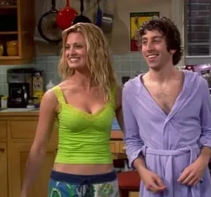 Mulher de top verde e homem de roupão roxo sorriem em uma cozinha. Um terceiro de robe xadrez à esquerda. Cena alegre.