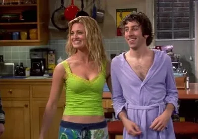 Mulher de top verde e homem de roupão roxo sorriem em uma cozinha. Um terceiro de robe xadrez à esquerda. Cena alegre.