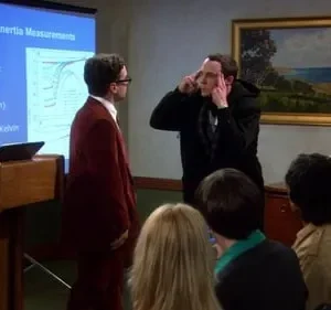 Dois homens, um de terno vermelho e outro com mãos nas têmporas, numa apresentação científica com plateia. Clima tenso.