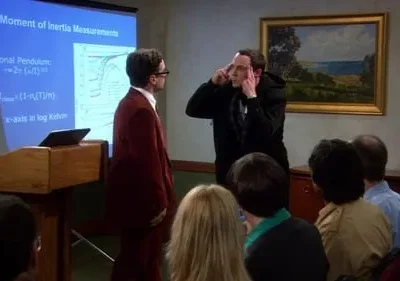 Dois homens, um de terno vermelho e outro com mãos nas têmporas, numa apresentação científica com plateia. Clima tenso.