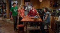 Quatro amigos em uma cozinha. Um homem de camiseta geek, uma mulher loira, um homem de roupão e um de colete jeans interagem casualmente.