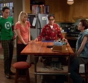Quatro amigos em uma cozinha. Um homem de camiseta geek, uma mulher loira, um homem de roupão e um de colete jeans interagem casualmente.